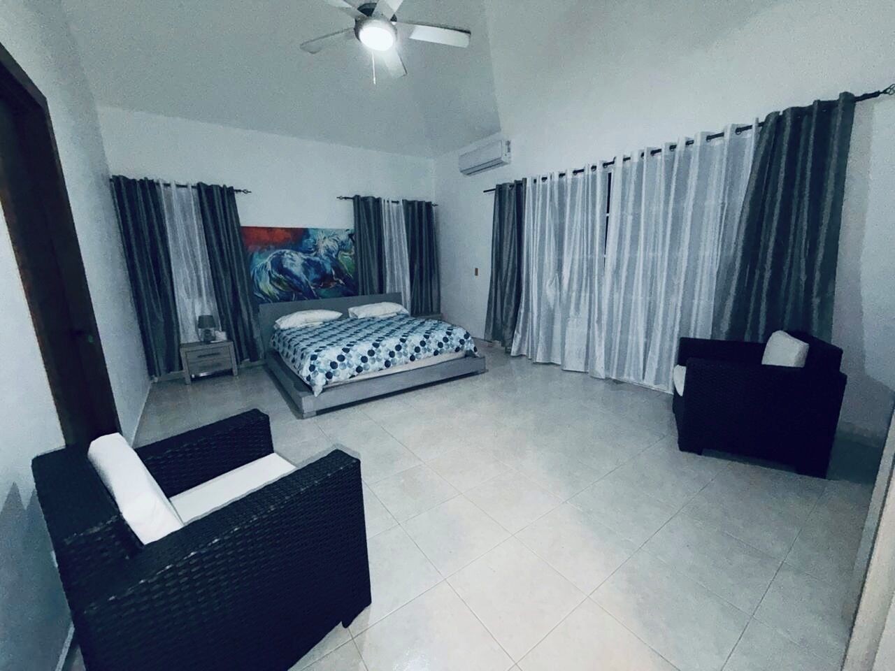 Un dormitorio con una cama, dos sofás negros, un ventilador de techo y un ventanal con cortinas.