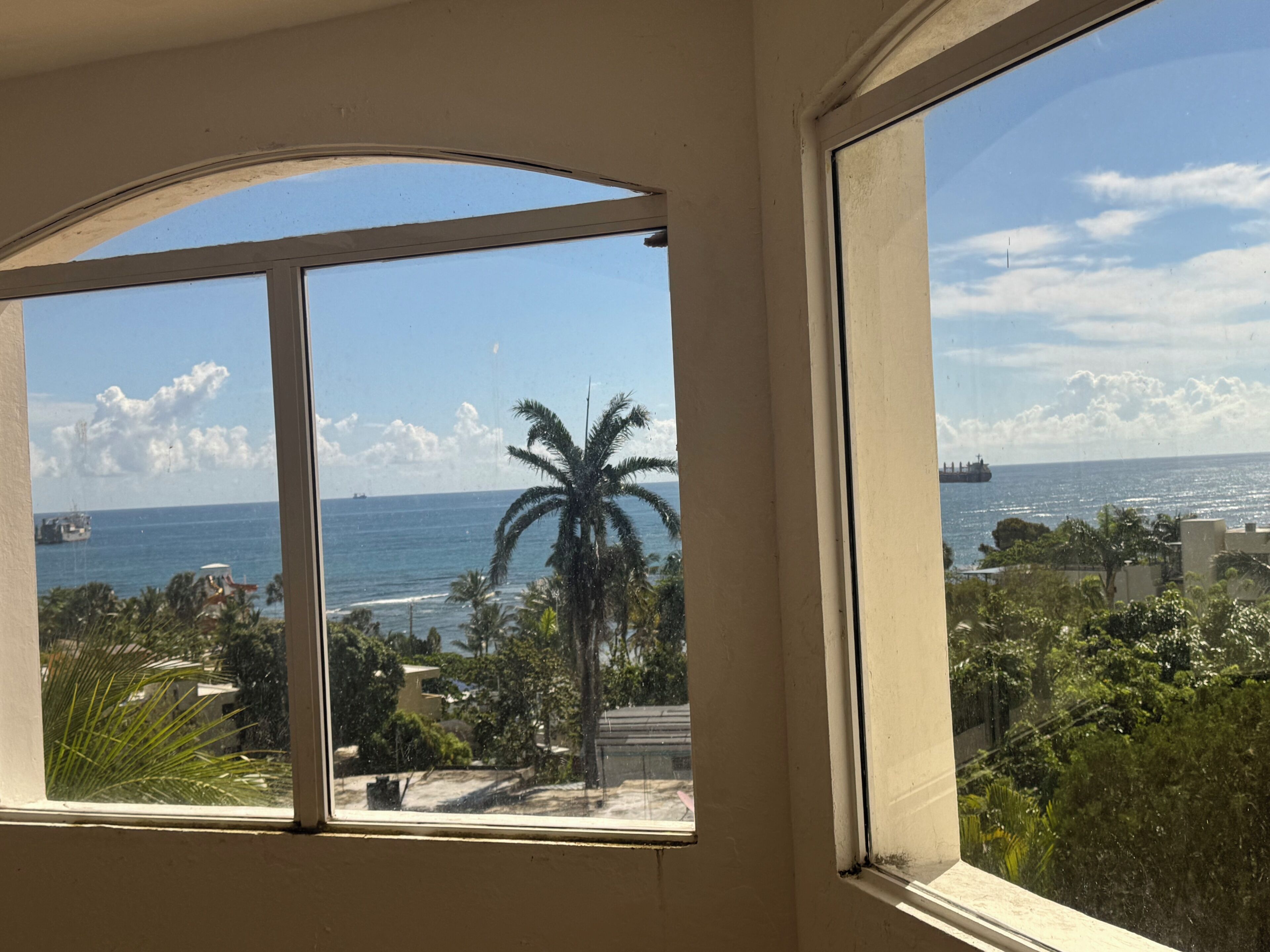 A room with a large window offering a view of the ocean, palm trees, and a distant ship.