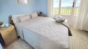 A bedroom with a bed, a nightstand, a framed picture, and a window with a view.