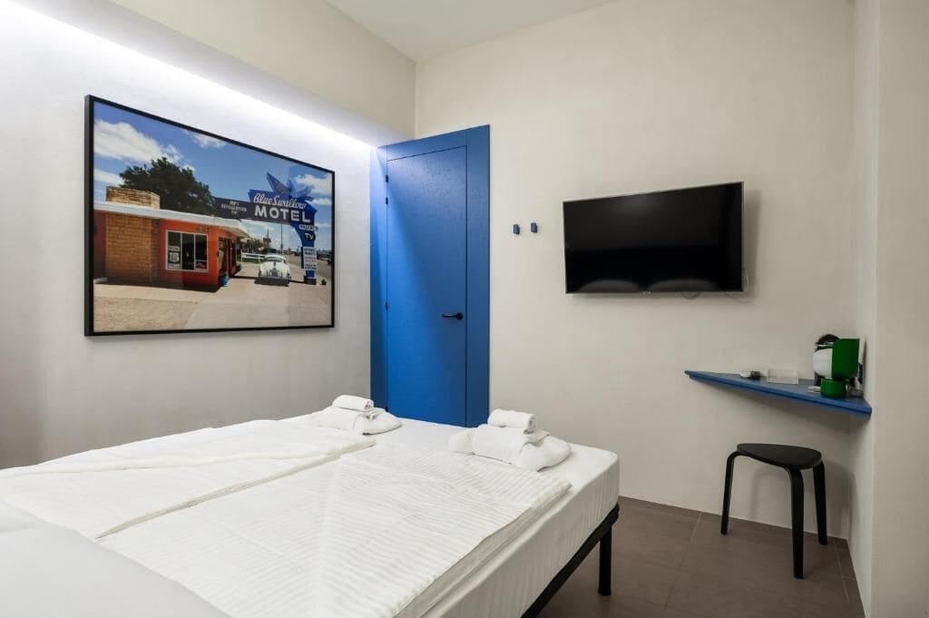 A hotel room with a bed, a blue door, a TV mounted on the wall, and a small stool.