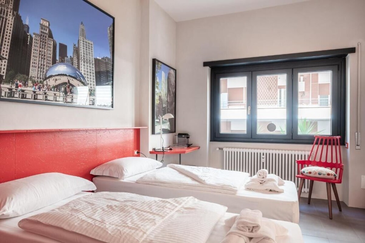 Habitación de hotel con dos camas, un cabecero rojo, una ventana con persianas, un radiador, una silla roja y un cuadro enmarcado de un paisaje urbano.