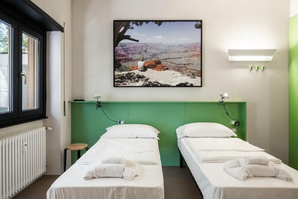 Habitación de hotel con dos camas, cabecera verde, ventana con persianas, un cuadro de un cañón y un taburete pequeño.