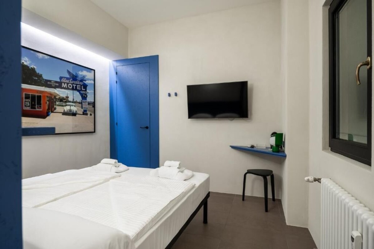 Una habitación de hotel con una cama, un televisor montado en la pared, una puerta azul y un taburete pequeño.