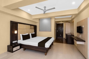 Quarto de hotel moderno com uma cama grande, ventoinha no teto, TV de tela plana e banheiro visível por uma porta aberta.