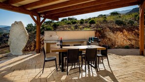 Een terras buiten, met een houten pergola, een stenen muur, een barbecue en een tafel met stoelen.