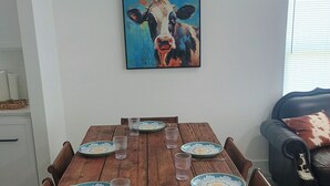 Ruokapöytä, jossa on lautasia ja laseja, seinällä lehmäaiheinen taulu ja taustalla sohva.