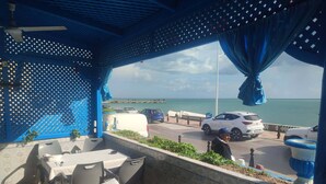 An outdoor seating area with tables and chairs, a view of the sea, and parked cars.