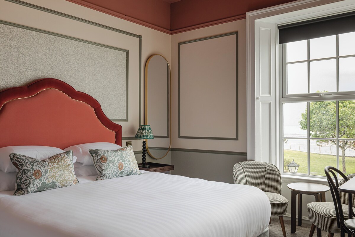 Een hotelkamer met een groot bed, een zithoek met een fauteuil en een tafel, een raam met uitzicht en een spiegel aan de muur.