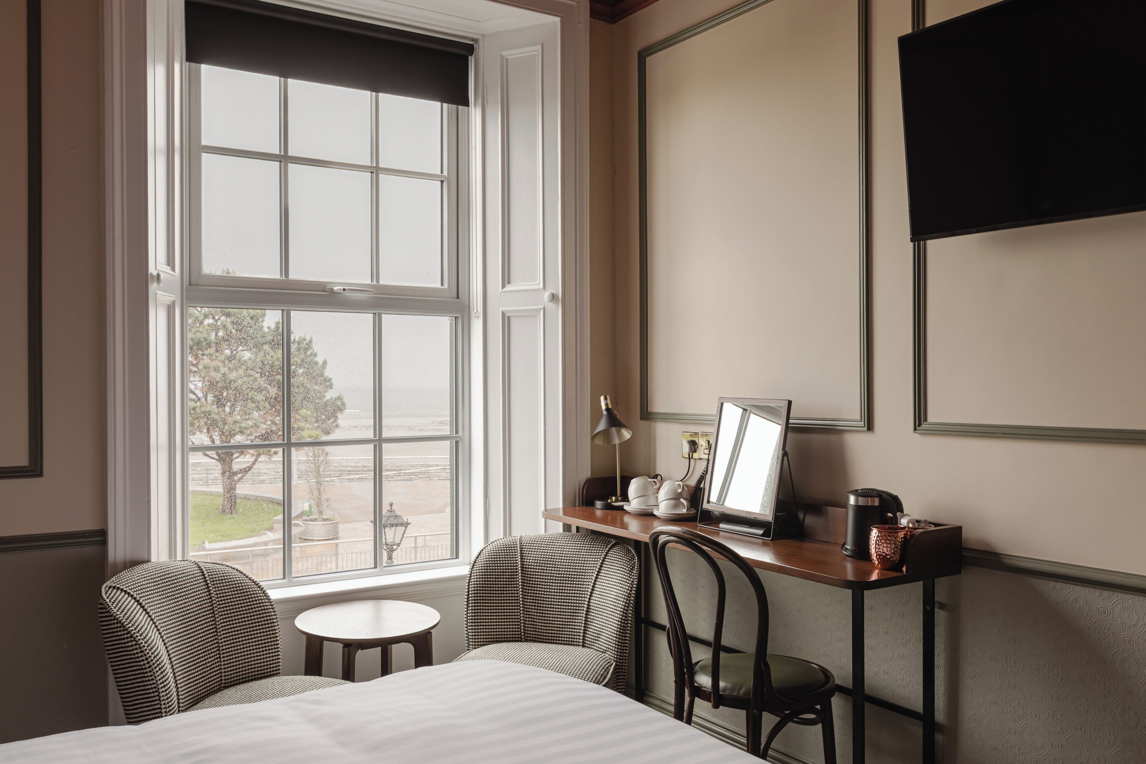 Habitación de hotel con un ventanal grande, un escritorio con computadora, dos sillas, una cama y un televisor en la pared.