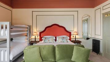 A hotel room with a bunk bed, a large bed with green upholstered headboard, two bedside tables with lamps, a large mirror, and a radiator.