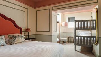 Habitación de hotel con una cama grande, una silla, una mesita, un espejo y una cama litera al fondo.