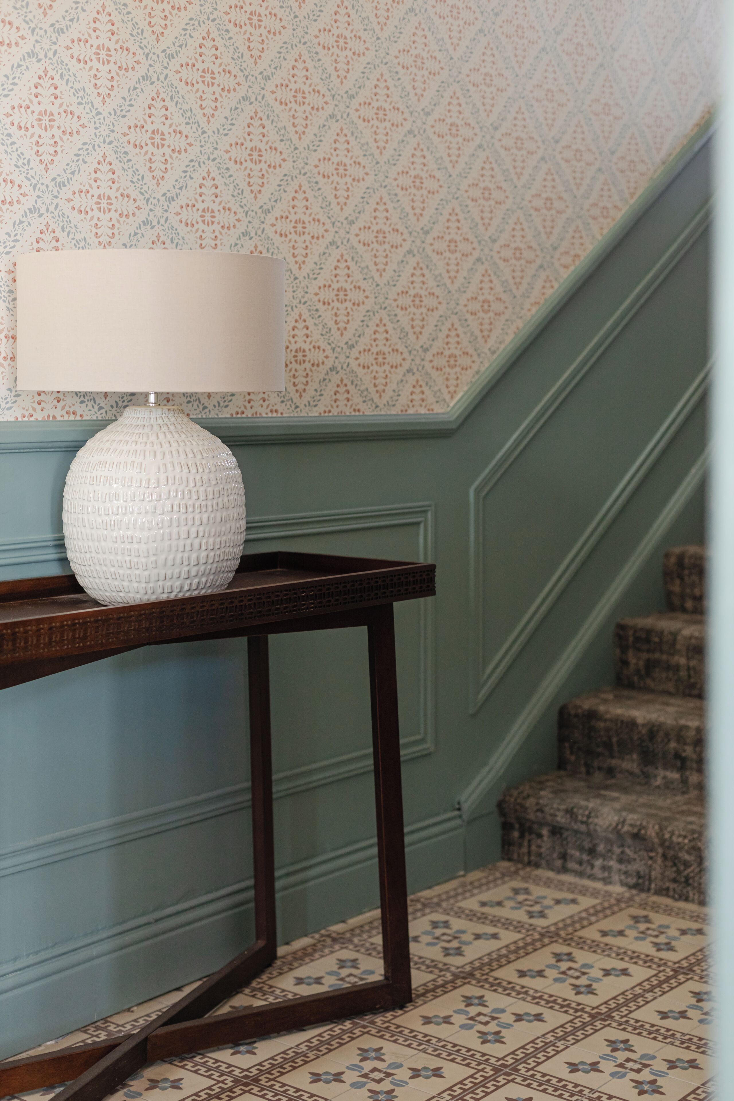 Un escalier recouvert d’une moquette à motifs et une lampe blanche posée sur une console en bois.