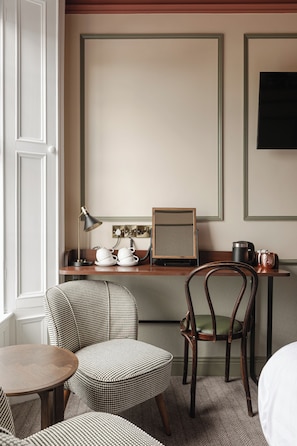 A hotel room with a round wooden table, a houndstooth patterned ottoman, a wooden chair, a desk with a lamp, a telephone, and a framed picture on the wall. - Avoca House (Newcastle)