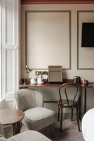 Habitación de hotel con una mesa redonda de madera, un taburete a cuadros de piedritas, una silla de madera, un escritorio con lámpara, un teléfono y un cuadro en la pared.