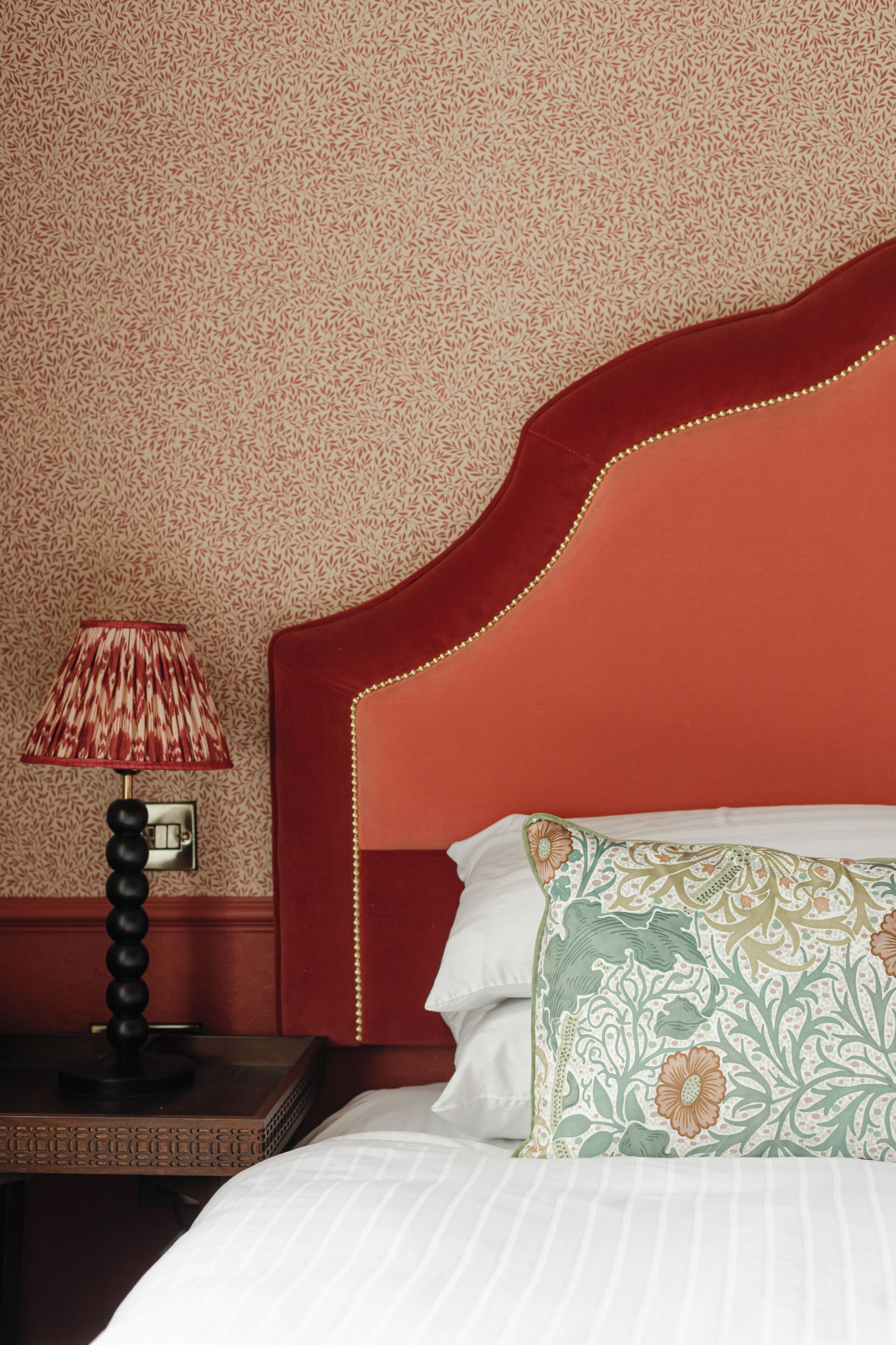 Una habitación de hotel con un cabecero rojo, una lámpara estampada y una almohada con motivos florales.