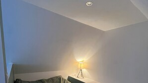 Une petite pièce à la lumière tamisée, avec un lit simple, une table de chevet et une lampe fixée au mur.