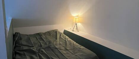 Um quarto pequeno e pouco iluminado, com uma cama de solteiro, uma mesa de cabeceira e uma luminária na parede.