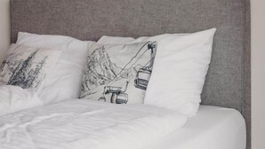 Una cama con cabecera gris y ropa de cama blanca, con dos almohadas que tienen ilustraciones de montañas y telesillas.