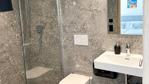 Un baño con ducha de vidrio, inodoro blanco, lavamanos con dispensador de jabón negro y un taburete de madera pequeño.