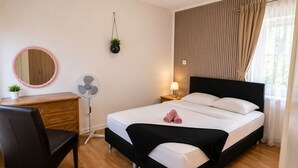 Une chambre à coucher avec un lit, un bureau, une chaise, un miroir, un ventilateur et une fenêtre avec des rideaux.