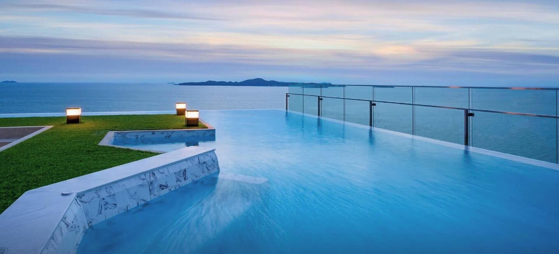A infinity pool with a glass barrier, a grassy area, and a view of the sea.