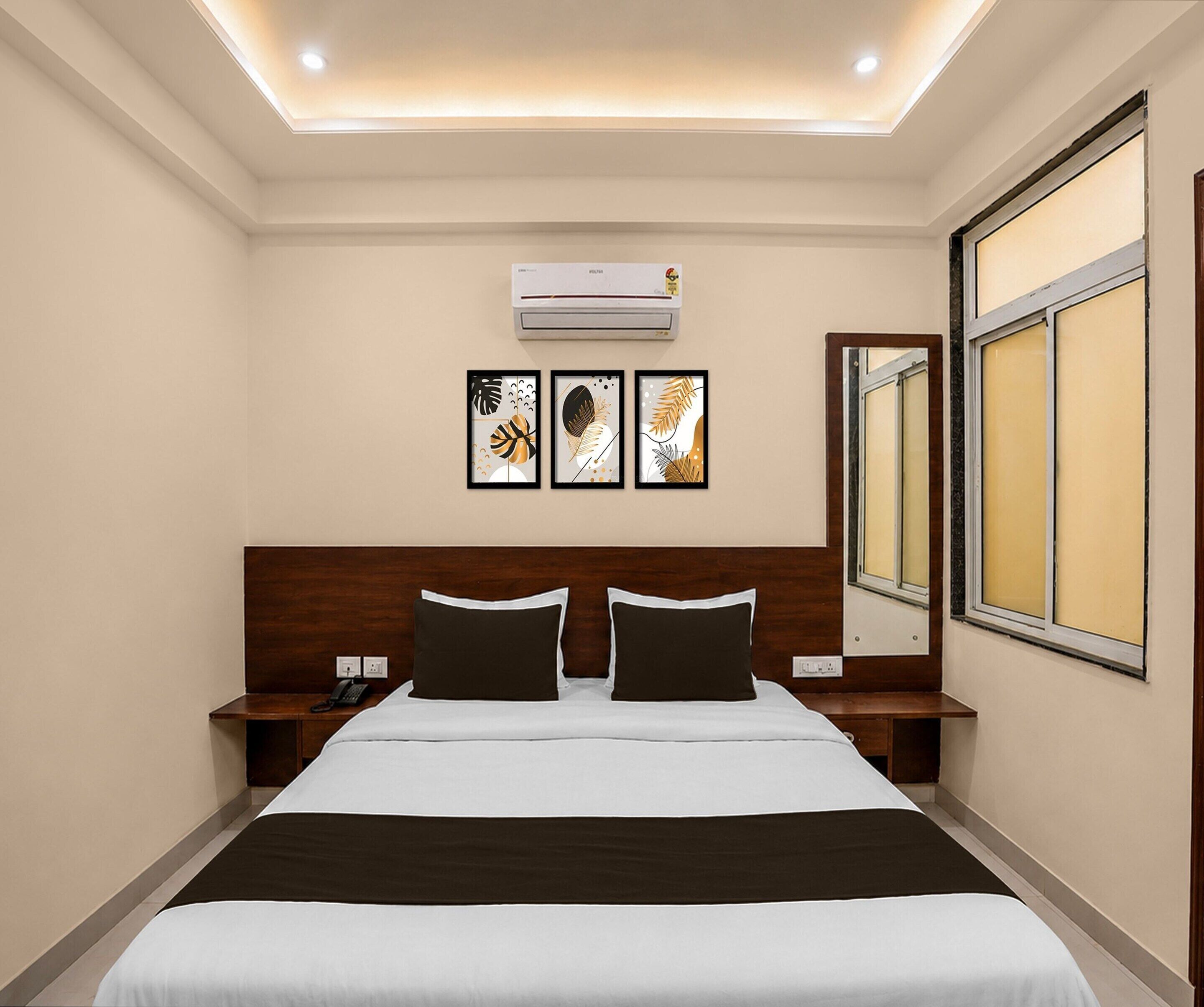 A hotel room with a bed, bedside tables, an air conditioning unit, a large window, and three framed artworks on the wall.