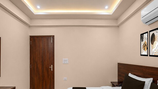 A hotel room with a wooden door, a bed with a white and brown bedspread, a nightstand, a bedside table, and an air conditioning unit on the wall.