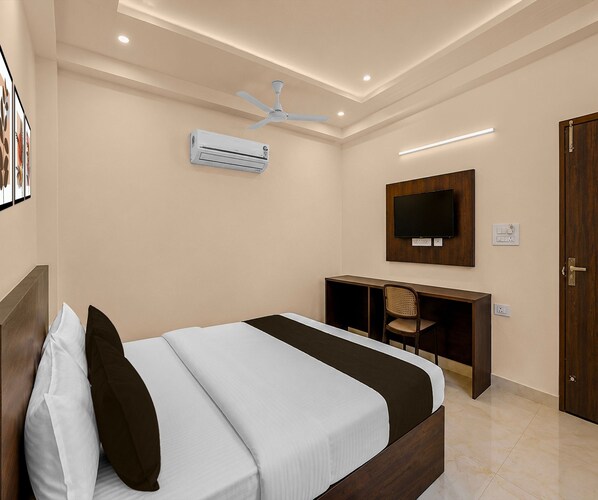 A hotel room with a bed, desk, chair, TV, air conditioning unit, and a door.