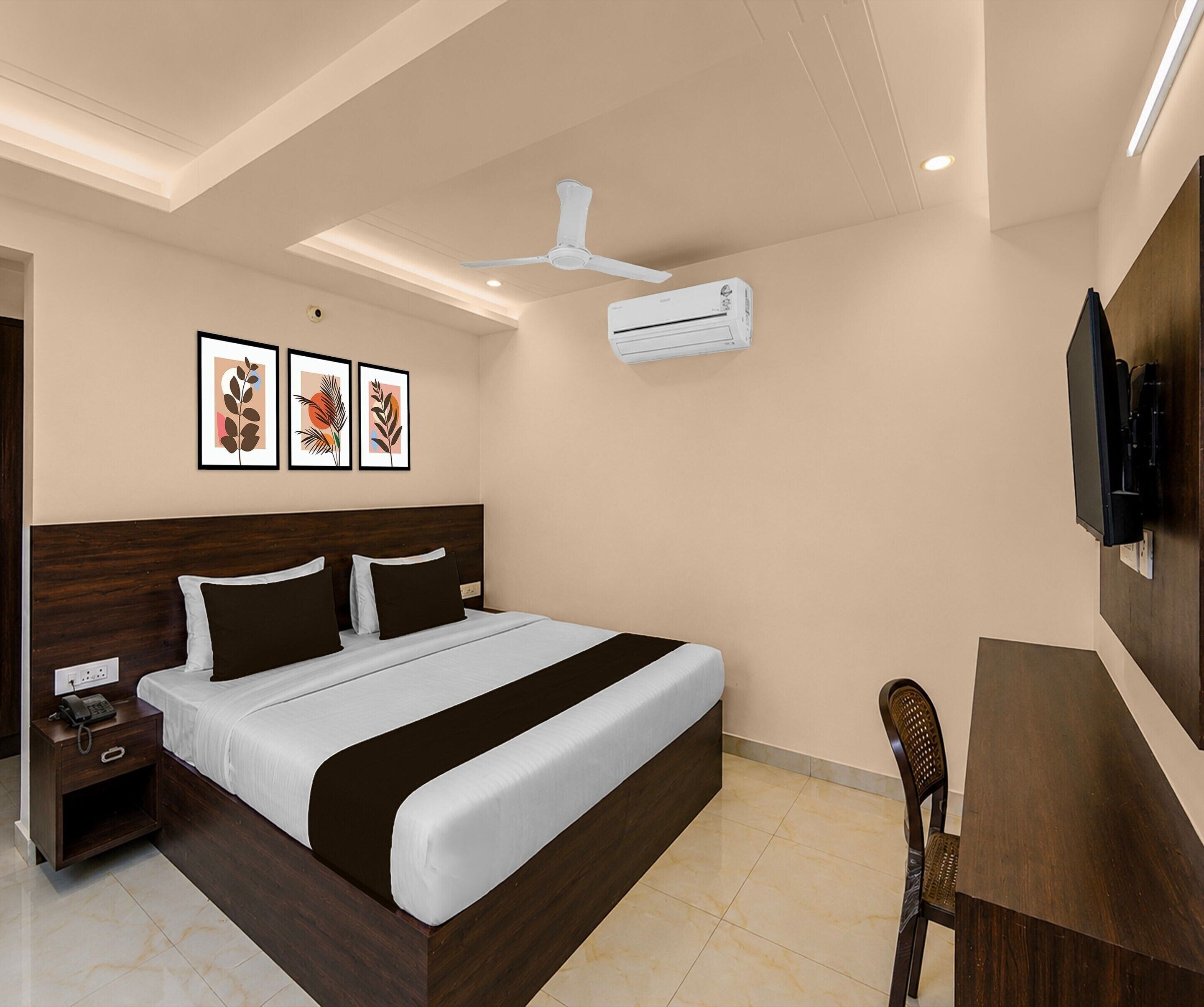 A hotel room with a bed, bedside table, phone, wall-mounted TV, ceiling fan, air conditioning unit, and framed artwork.