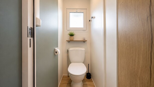 A narrow bathroom with a toilet, a small window above the toilet, a shelf with a plant, and a door on the left. - Le Cocon du Mar Estang - Terrace, air conditioning, sleeps 8 (Canet-en-Roussillon)