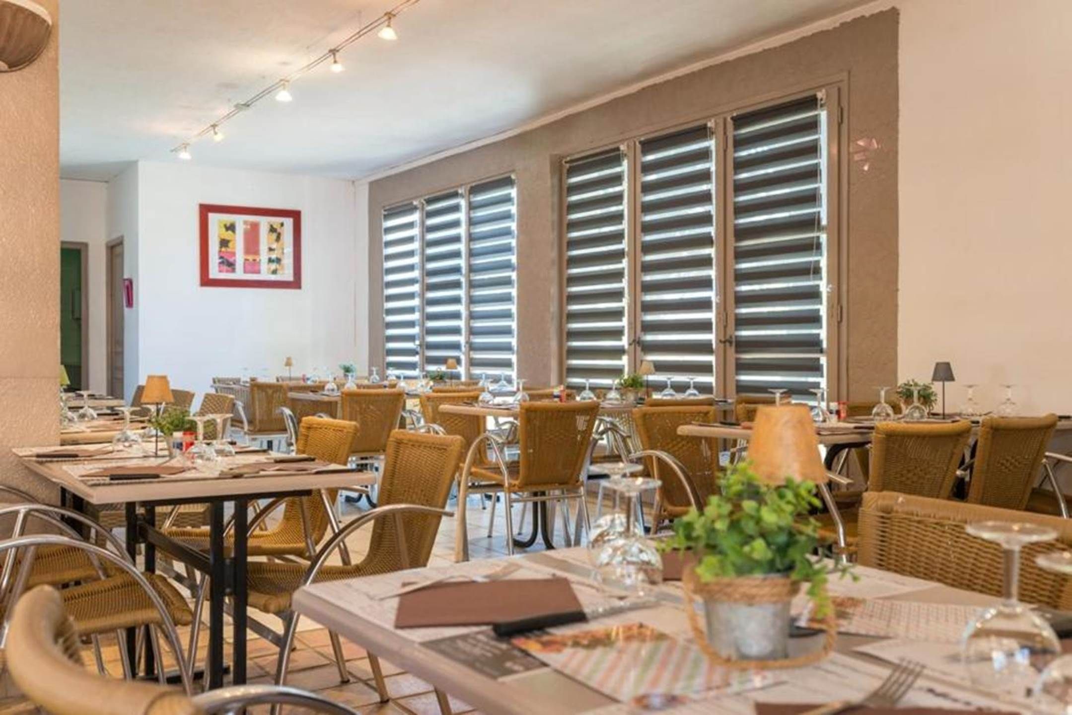 Un comedor con mesas y sillas, ventanas amplias con persianas y un cuadro en la pared.
