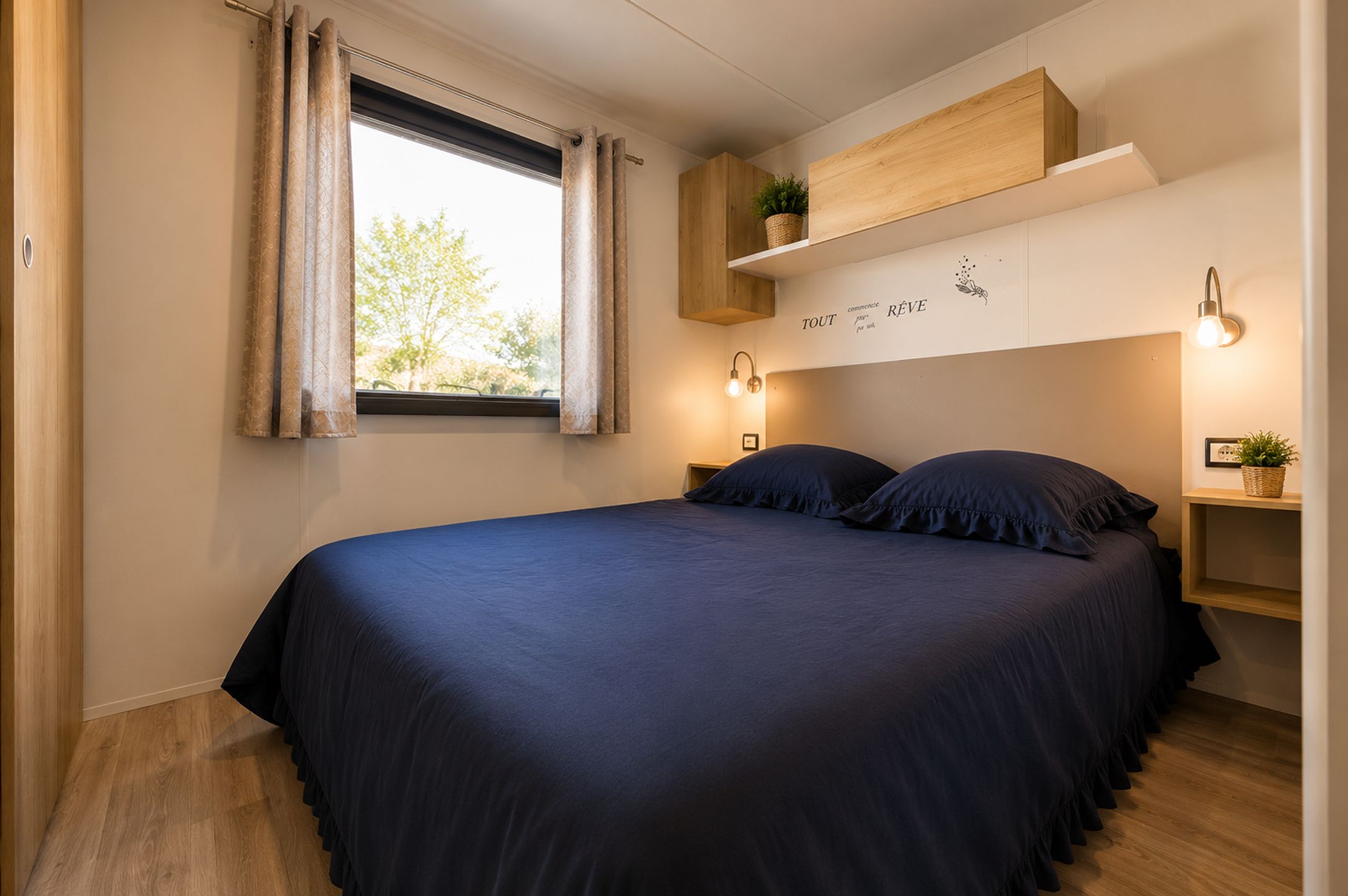 Ein Schlafzimmer mit Bett, Vorhängen, einem Fenster, einem Regal mit Pflanze und Wandleuchten.