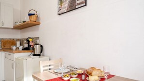Un comedor con una mesa preparada para dos, una cesta en un estante y una zona de cocina con un refrigerador y una tetera.
