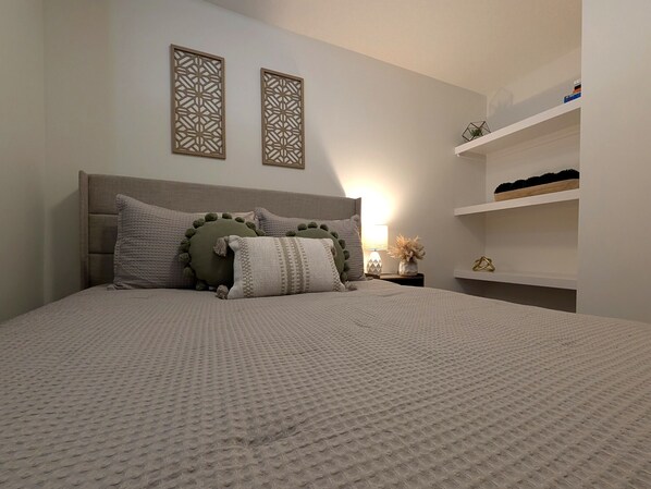 A neatly arranged bedroom with a bed, pillows, a nightstand, a wall-mounted shelf, and decorative wall panels.