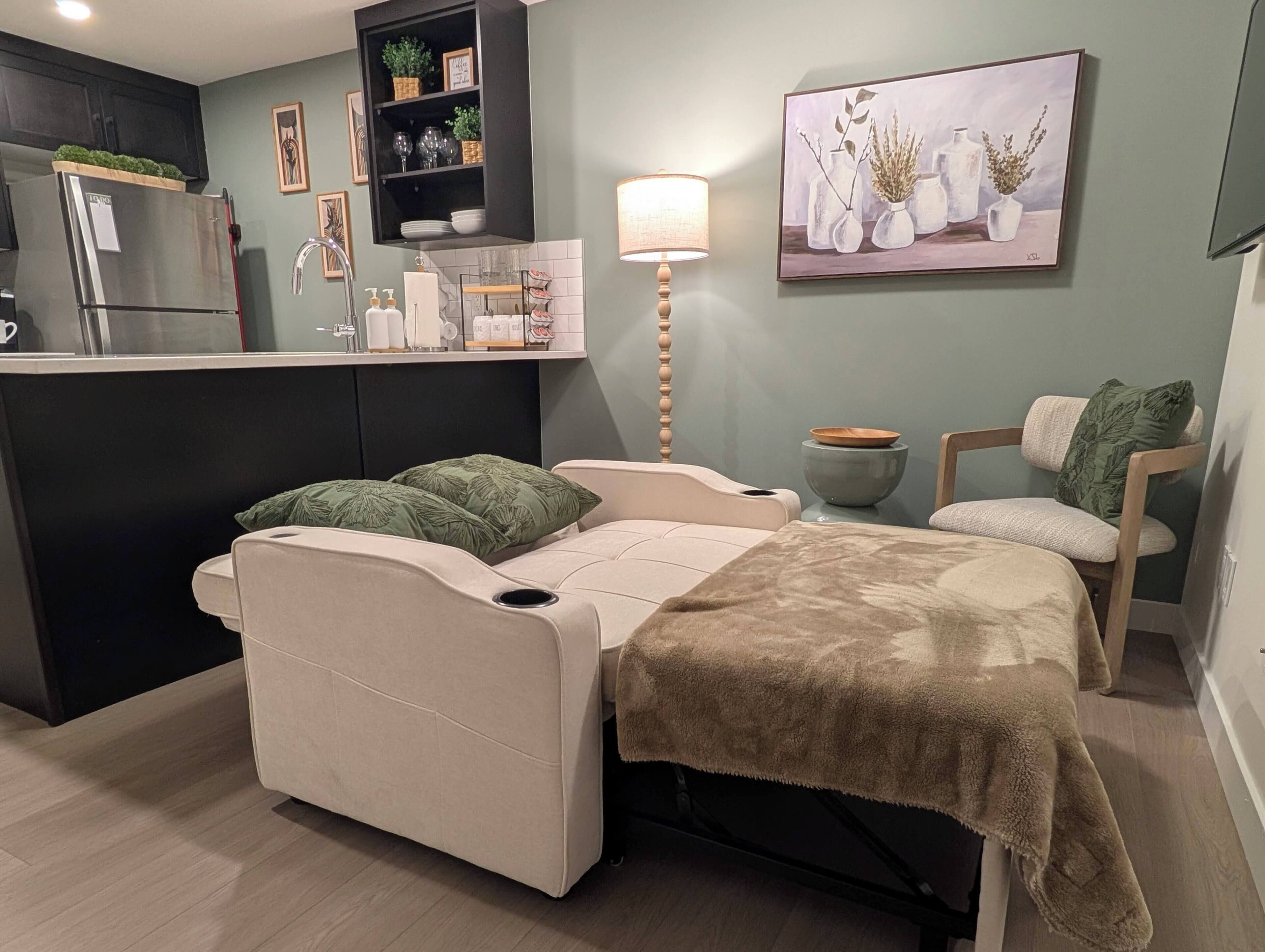 Un espacio habitable compacto con kitchenette, una cama con cabecera, una silla y un cuadro en la pared.
