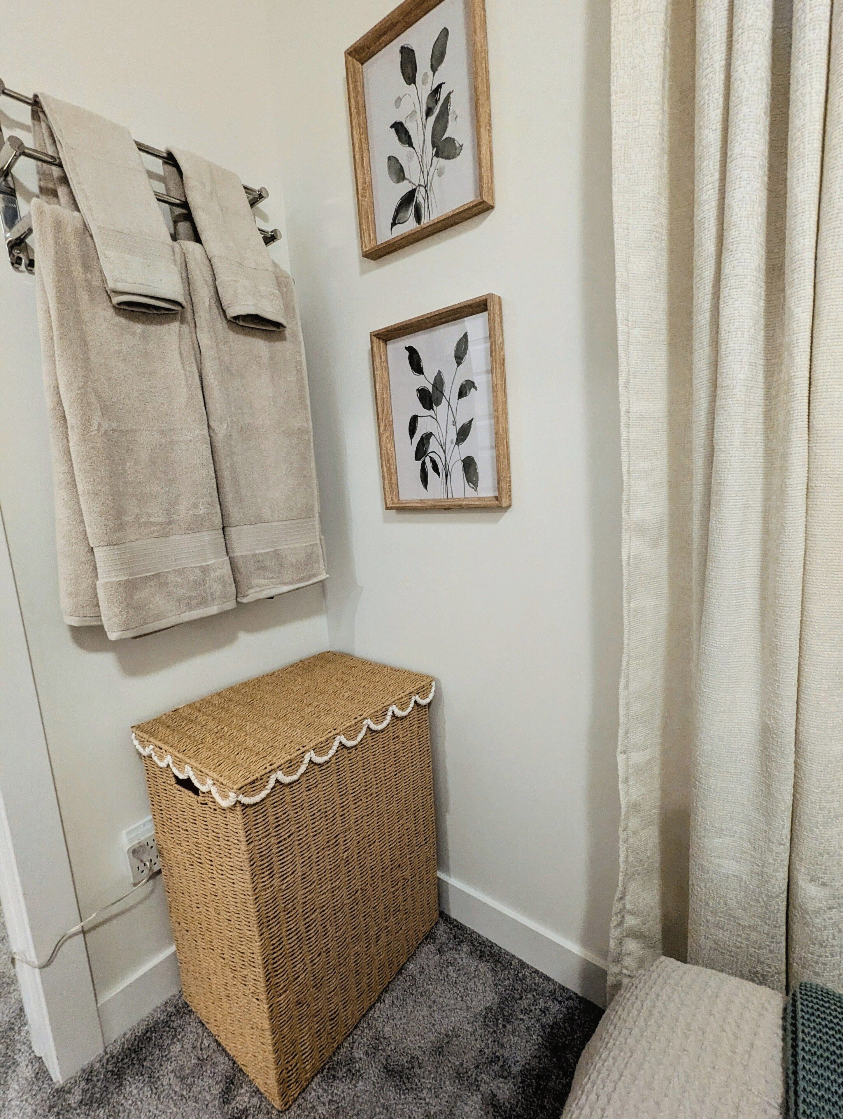 Een badkamer met een handdoekrek met twee handdoeken, een plant in een lijst aan de muur, een geweven mand en een gordijn.