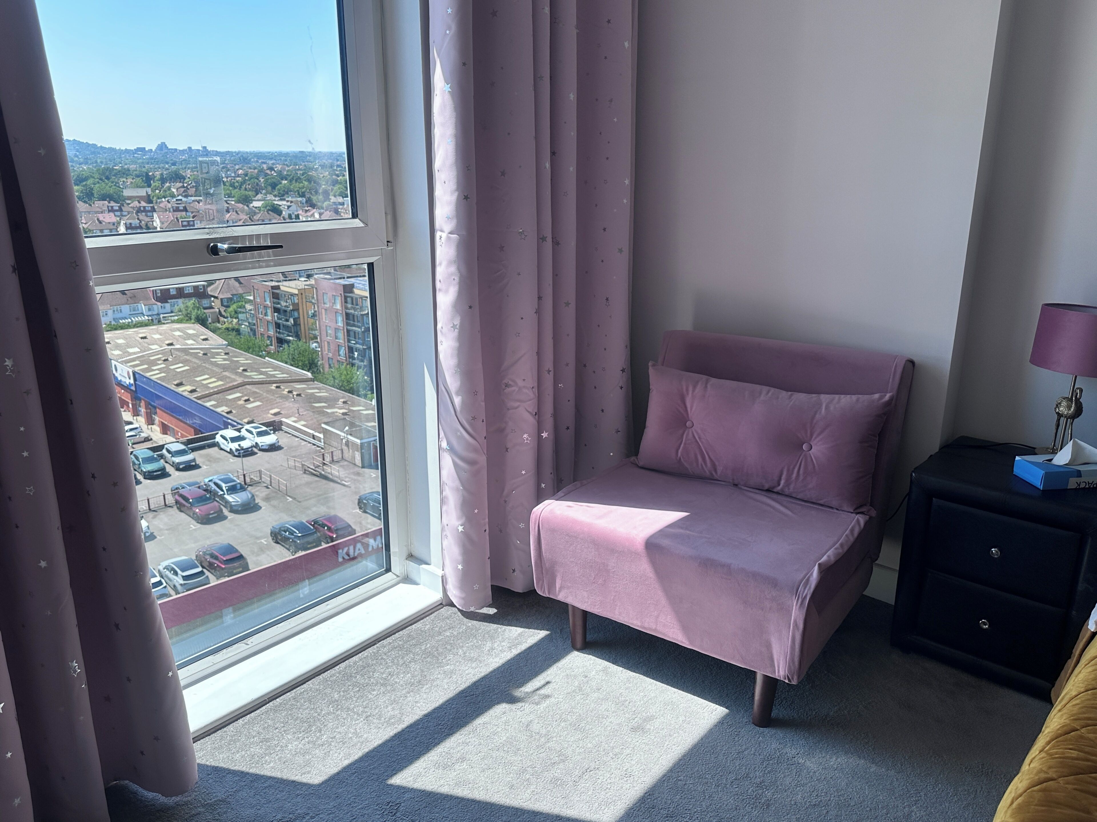 Ein Schlafzimmer mit Bett, Nachttisch, Lampe, einem Fenster mit Vorhängen und Blick auf einen Parkplatz und Gebäude.