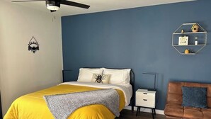 Ein Schlafzimmer mit einem Bett, einem Nachttisch, einem Sessel, einem Deckenventilator und einem Wandregal.
