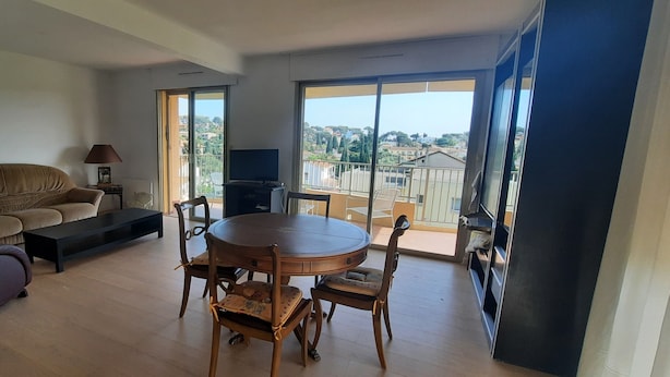 Uma sala de estar com sofá, mesa de centro, mesa de jantar com cadeiras, televisão e varanda com vista.