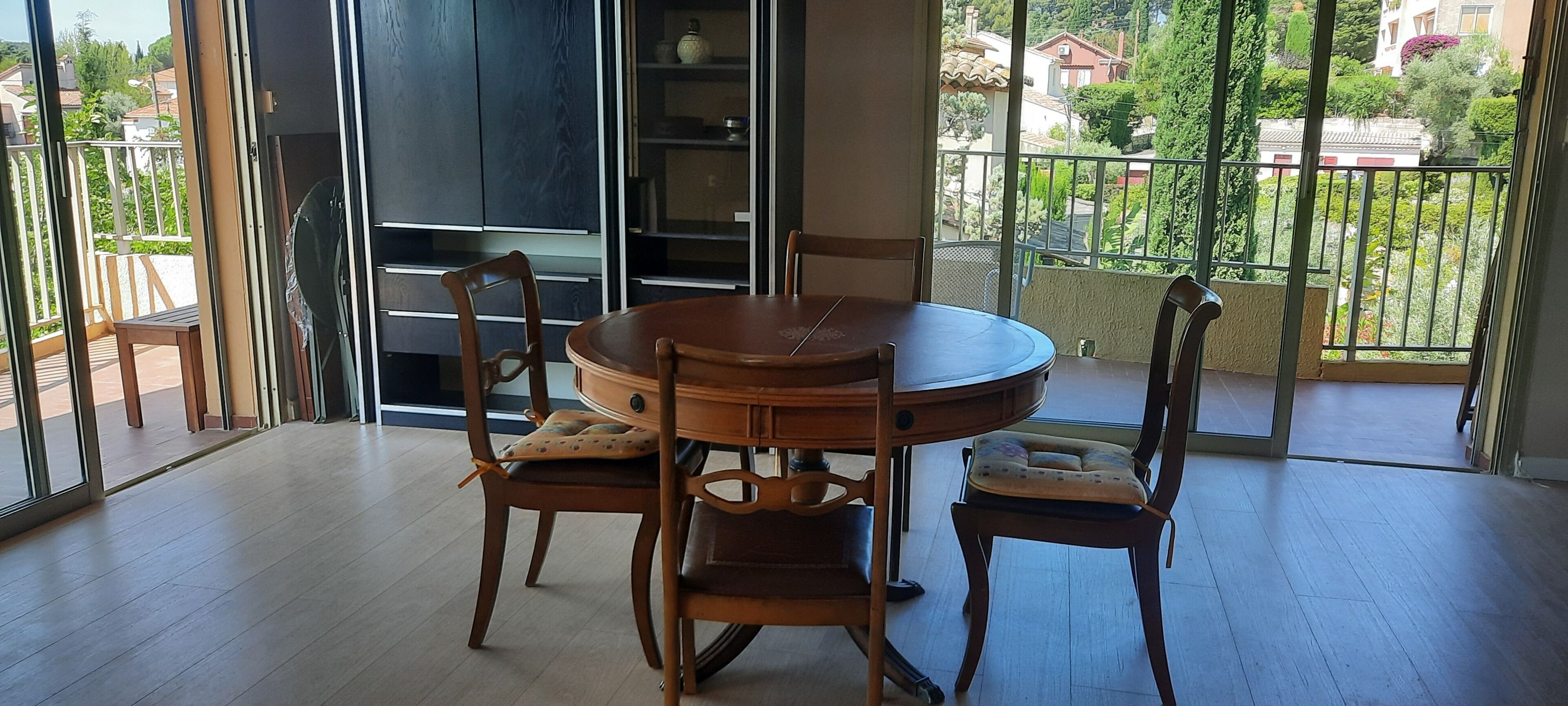 A dining area with a round table and four wooden chairs, a bookshelf, and a view of a balcony with greenery.
