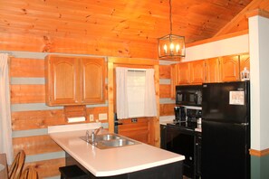 A kitchen with wooden cabinets, a stainless steel sink, a black refrigerator, and a hanging light fixture. - Mountains 6 mins to attractions + 2 king beds + hot tub + fire pit + pool access (Sevierville)