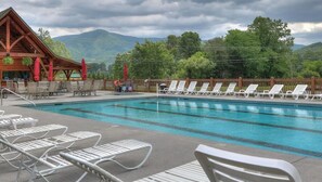 A pool area with lounge chairs, a wooden building with a sloped roof, and a mountainous backdrop. - Mountain getaway 6 mins to attractions + 2 king beds + hot tub + fire pit (Sevierville)