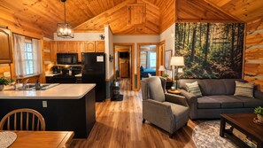 A cabin interior with a kitchen, dining area, and living room, featuring wooden floors and a forest-themed wall mural. - Mountain getaway 6 mins to attractions + 2 king beds + hot tub + fire pit (Sevierville)