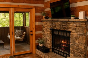 A cozy room with a stone fireplace, a television mounted above it, and a wooden door leading to an outdoor area with a swing chair. - Mountain getaway 6 mins to attractions + 2 king beds + hot tub + fire pit (Sevierville)