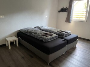 Een eenpersoonsbed met een zwarte matras en grijze beddengoed, een klein wit nachtkastje, een wandkapstok en een raam met gordijnen.