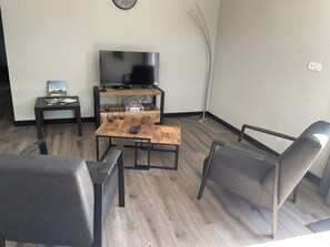 Een woonkamer met een televisie, een salontafel, twee stoelen, een bijzettafeltje met een boek en een vloerlamp.