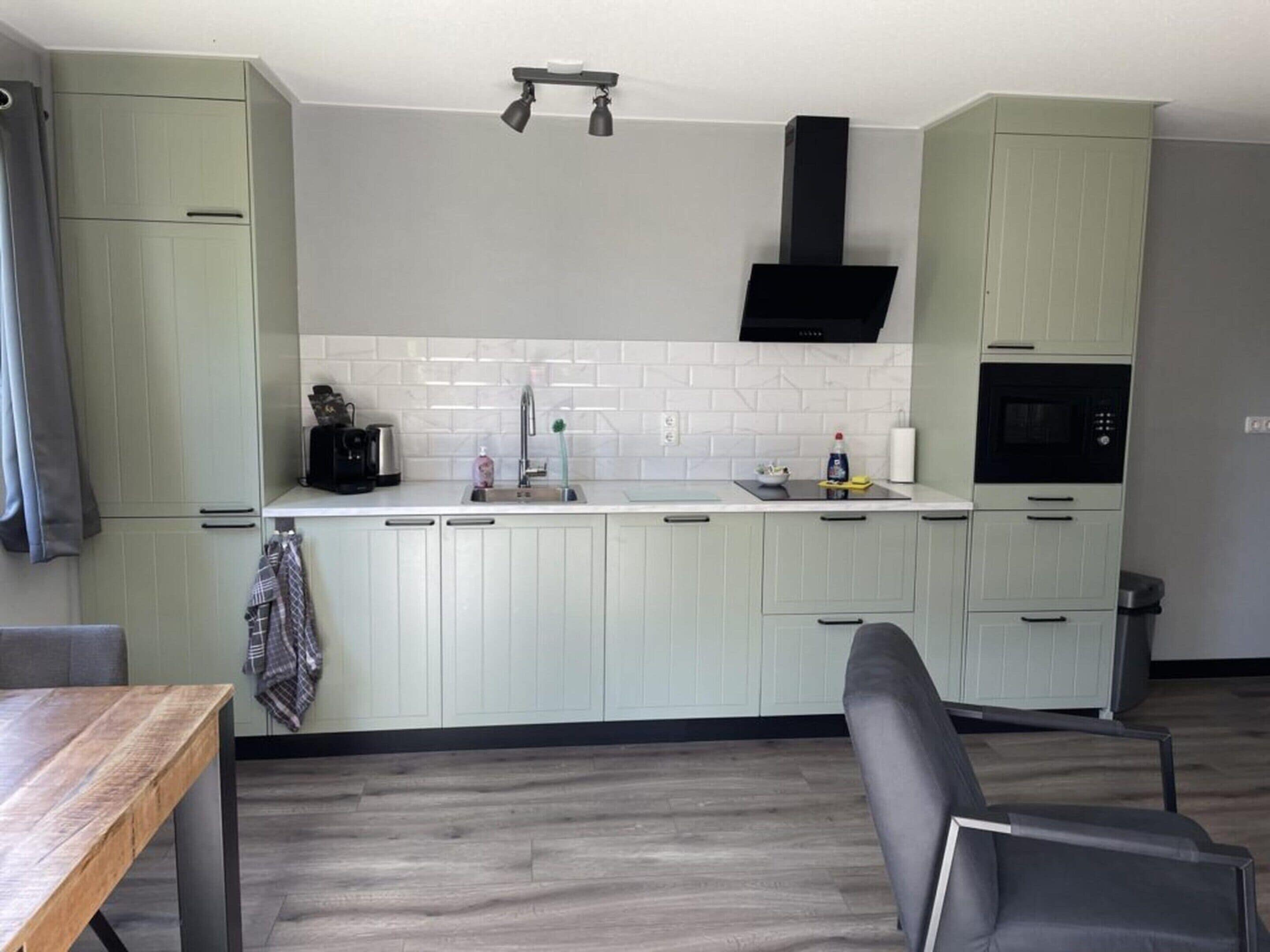A modern kitchen with light green cabinets, a white tiled backsplash, a black range hood, and a built-in oven.