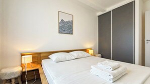 Una habitación de hotel con cama, mesita de noche, lámpara, silla, una obra de arte en la pared y armario.