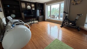 A living room with a television, exercise bike, and a large window with a view of trees. - 4BR Calgary Home | Ping Pong + Parking (Calgary)