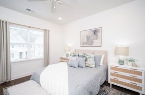 A bedroom with a bed, bedside tables, a ceiling fan, a window with curtains, and a wall-mounted abstract painting.